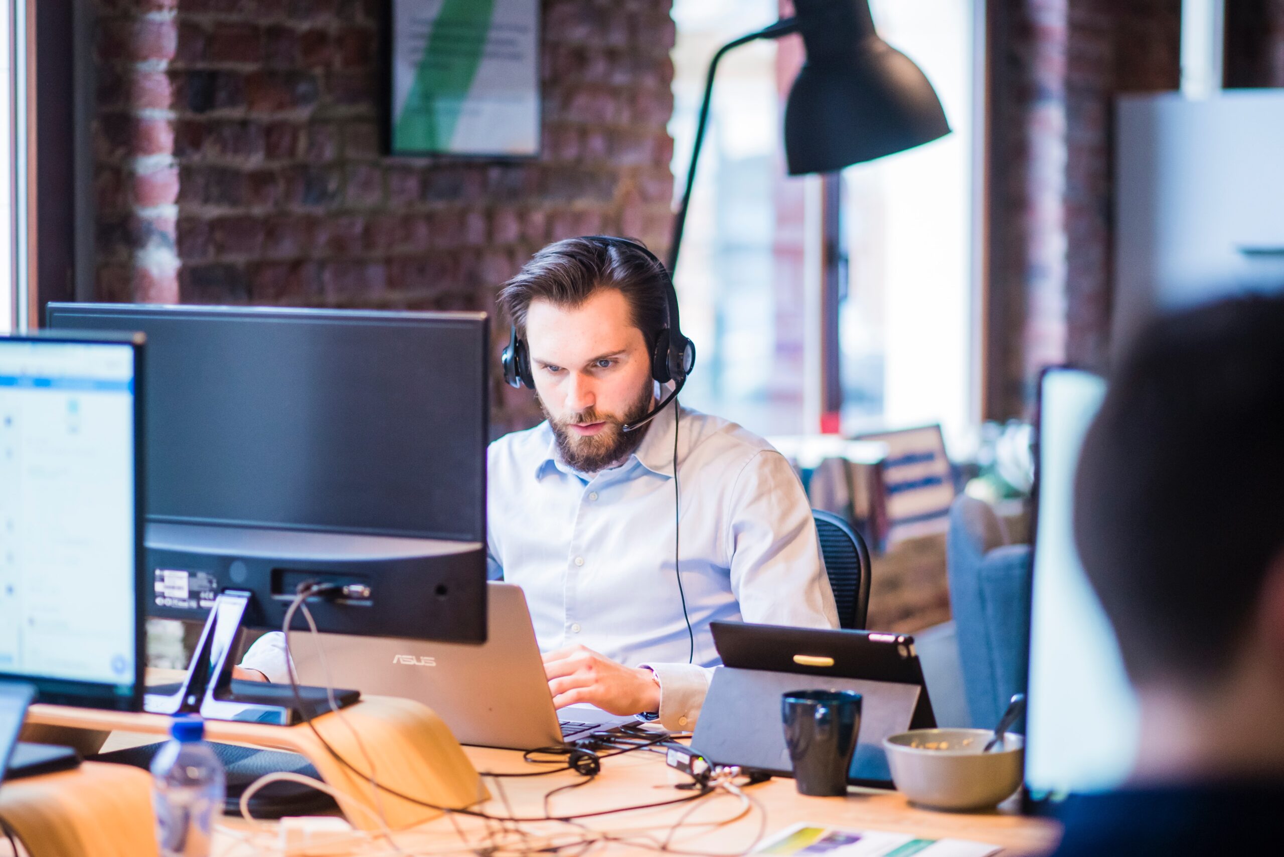Obrazek przedstawia mężczyznę siedzącego za biurkiem ze słuchawkami na uszach pracującego na laptopie